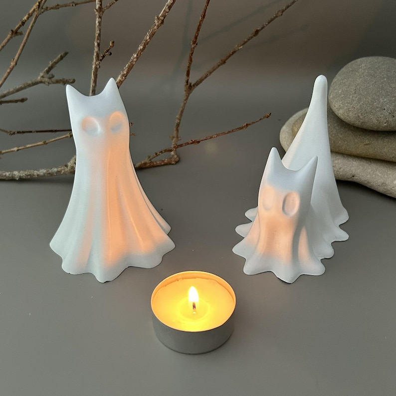 May include: Two white ghost-shaped figurines with smooth, flowing forms and hollow eye sockets, positioned next to a small, round metal tea light candle holder containing a lit candle with a warm, glowing yellow flame.