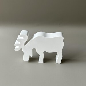 May include: A solid white, flat-surfaced moose figurine standing in profile. The object features a stylized, smooth silhouette with a distinct antler shape, a curved back, and four legs, crafted as a singular, continuous piece of white material.
