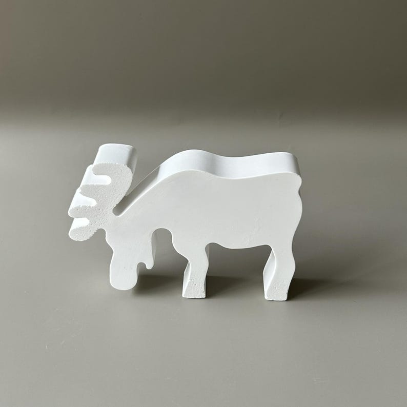May include: A solid white, flat-surfaced moose figurine standing in profile. The object features a stylized, smooth silhouette with a distinct antler shape, a curved back, and four legs, crafted as a singular, continuous piece of white material.