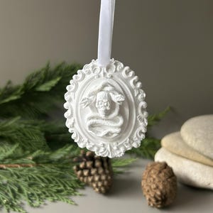 K&ouml;nnte beinhalten: Ein wei&szlig;es, ovales H&auml;ngeornament mit einem Relief eines Sch&auml;dels, der von einer Schlange umschlungen ist, eingefasst in einen verzierten Rahmen. Das Objekt ist an einem wei&szlig;en Band befestigt und zeigt detaillierte Verzierungen am Rand.