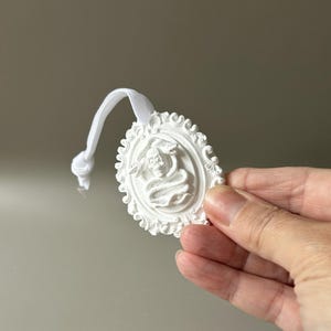 K&ouml;nnte beinhalten: Ein wei&szlig;es, ovales Ornament mit einem Reliefdesign aus einem Totenkopf, der von einer Schlange umschlungen ist, umgeben von einem verzierten Rand. Das St&uuml;ck ist an einer wei&szlig;en elastischen Kordel mit einem Knoten befestigt, die als Aufh&auml;ngeschlaufe dient.
