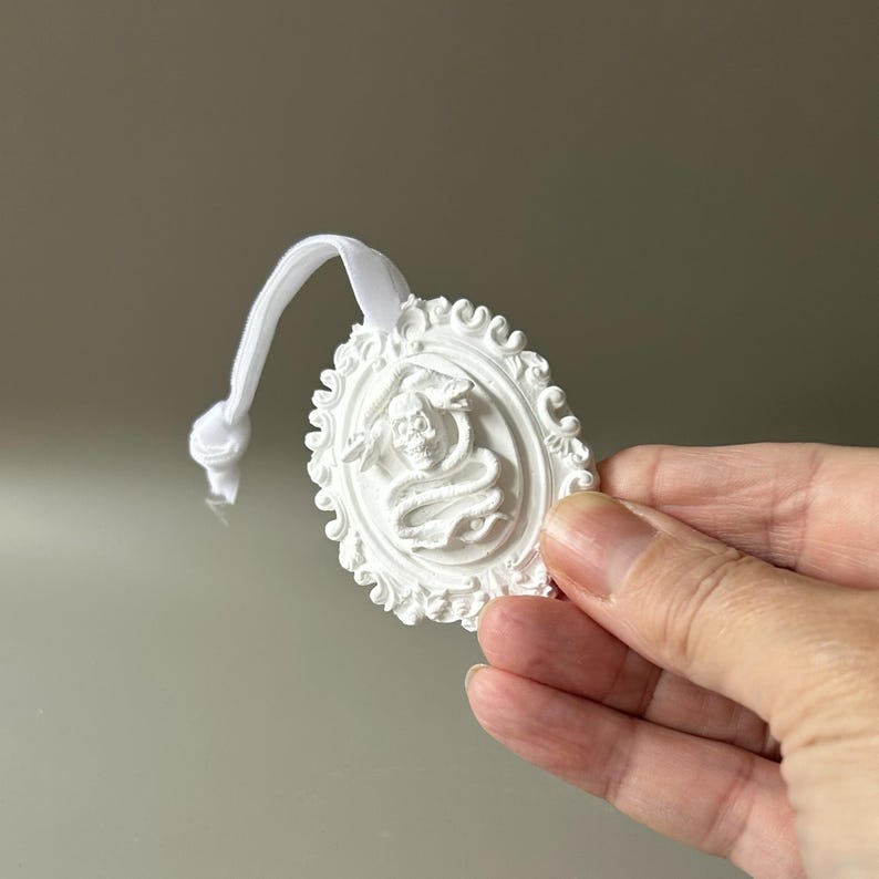 K&ouml;nnte beinhalten: Ein wei&szlig;es, ovales Ornament mit einem Reliefdesign aus einem Totenkopf, der von einer Schlange umschlungen ist, umgeben von einem verzierten Rand. Das St&uuml;ck ist an einer wei&szlig;en elastischen Kordel mit einem Knoten befestigt, die als Aufh&auml;ngeschlaufe dient.