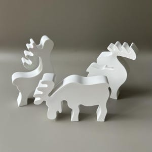 May include: Three white, matte-finished, stylized moose figurines. Two of the moose are standing upright, while one is positioned in the foreground in a grazing posture. Each figure features a smooth, solid surface and distinct, simplified antler shapes.