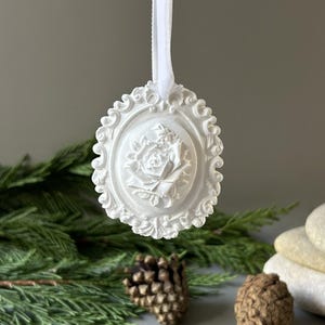 K&ouml;nnte beinhalten: Ein wei&szlig;es, ovales H&auml;ngeornament mit einem detaillierten Relief einer bl&uuml;henden Rose in der Mitte. Das Ornament ist von einem verzierten, schn&ouml;rkelartigen Rand umgeben und wird an einem d&uuml;nnen wei&szlig;en Band aufgeh&auml;ngt, das oben am Rahmen befestigt ist.