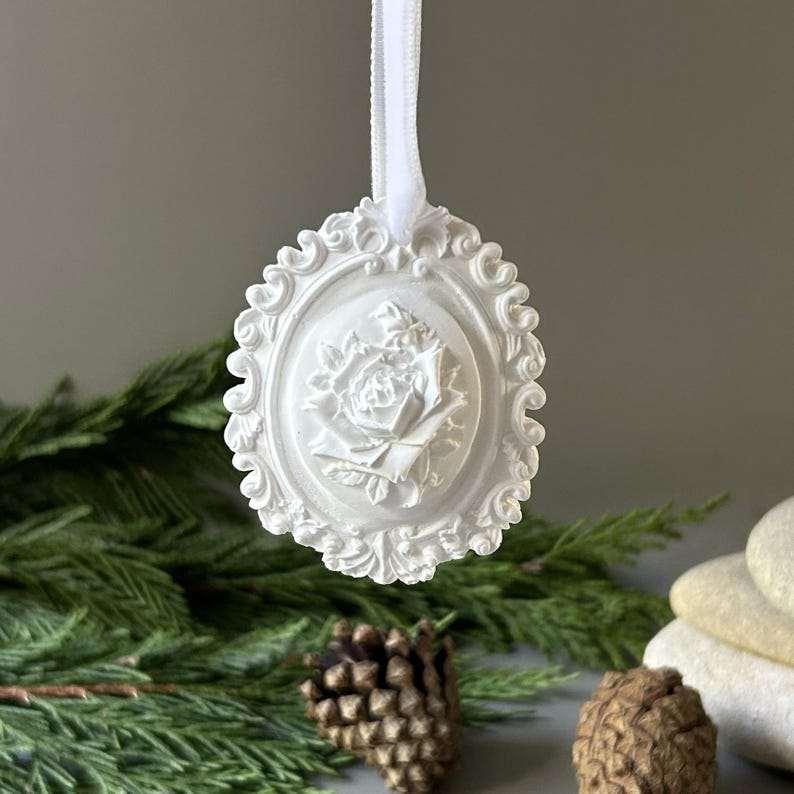 K&ouml;nnte beinhalten: Ein wei&szlig;es, ovales H&auml;ngeornament mit einem detaillierten Relief einer bl&uuml;henden Rose in der Mitte. Das Ornament ist von einem verzierten, schn&ouml;rkelartigen Rand umgeben und wird an einem d&uuml;nnen wei&szlig;en Band aufgeh&auml;ngt, das oben am Rahmen befestigt ist.