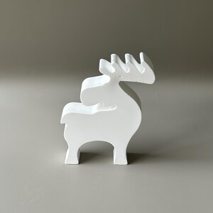 May include: A solid white, matte-finished figurine shaped like a stylized reindeer or moose. The object features a smooth, minimalist silhouette with a distinct head, a curved back, four sturdy legs, and a prominent set of antlers extending upwards.