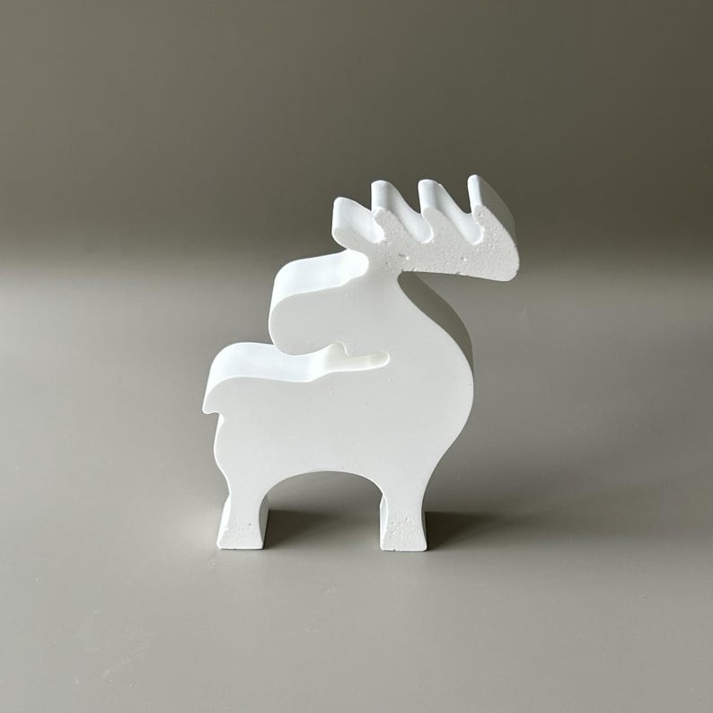 May include: A solid white, matte-finished figurine shaped like a stylized reindeer or moose. The object features a smooth, minimalist silhouette with a distinct head, a curved back, four sturdy legs, and a prominent set of antlers extending upwards.