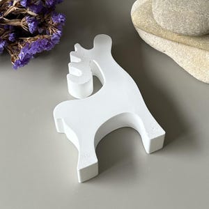 May include: A solid white, matte-finished figurine shaped like a stylized reindeer. The object features a smooth, minimalist silhouette with a curved neck, a distinct antler structure, and four legs, standing upright as a decorative tabletop ornament.