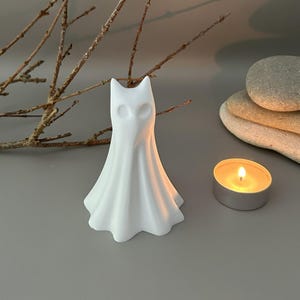 May include: A white, matte-finished ghost figurine with a pointed top and hollowed-out eye sockets. The figure has a draped, flowing shape that flares at the base, resembling a sheet-covered form. It stands upright as a singular decorative object.