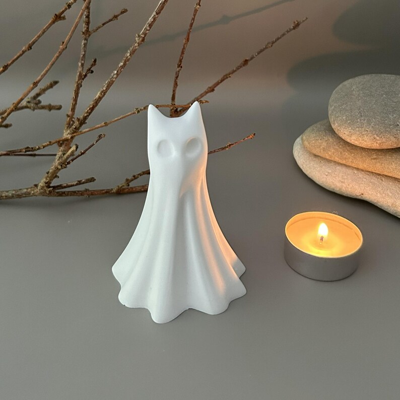 May include: A white, matte-finished ghost figurine with a pointed top and hollowed-out eye sockets. The figure has a draped, flowing shape that flares at the base, resembling a sheet-covered form. It stands upright as a singular decorative object.