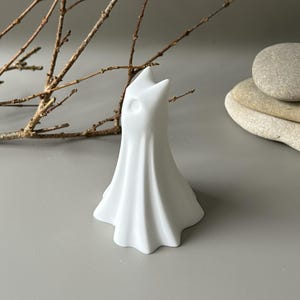 May include: A matte white ceramic figurine shaped like a draped ghost with a pointed top and a single circular eye indentation. The object has a flared, pleated base that mimics the appearance of flowing fabric, standing upright as a solid decorative piece.