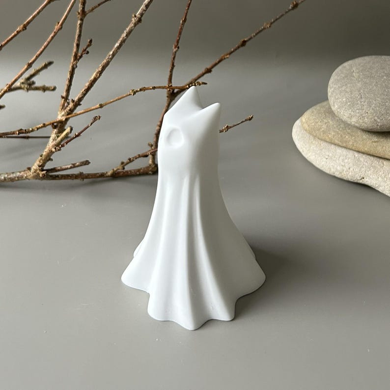 May include: A matte white ceramic figurine shaped like a draped ghost with a pointed top and a single circular eye indentation. The object has a flared, pleated base that mimics the appearance of flowing fabric, standing upright as a solid decorative piece.