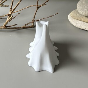 May include: A matte white ceramic object featuring a series of three connected, fluid, ghost-like forms with smooth, draped edges. The structure has a central opening at the top and a wide, scalloped base, presenting a clean, solid white finish.