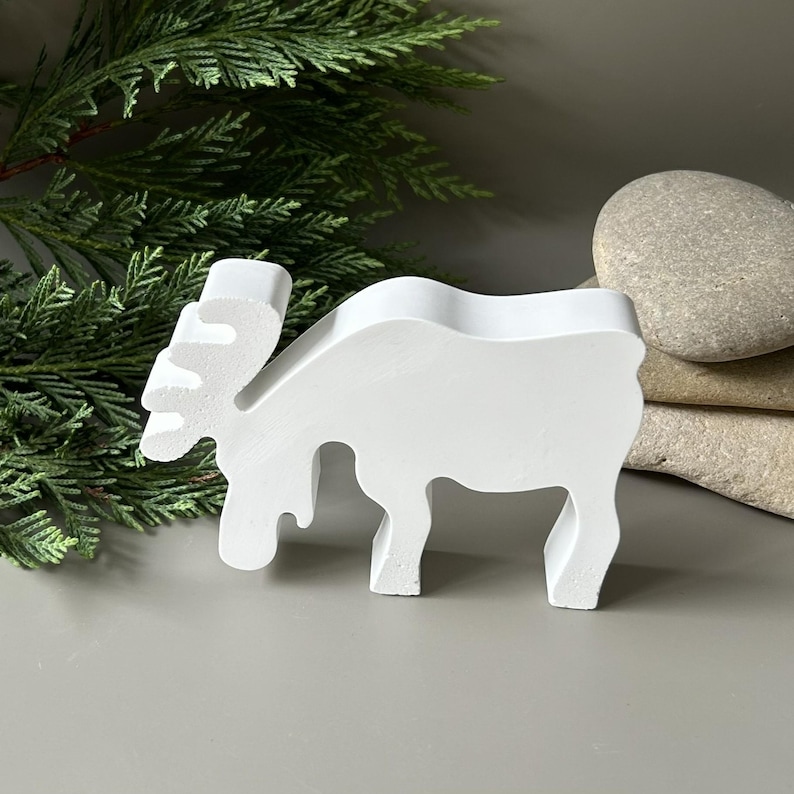 May include: A solid white, flat-cut figurine shaped like a moose in profile. The object features a smooth, matte finish with a distinct silhouette showing the animal's head, antlers, and four legs standing upright on a flat surface.