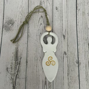 May include: White ceramic goddess pendant with a gold triple spiral design. The pendant is suspended by a green cord and a wooden bead. The goddess figure has outstretched arms and a stylized body.