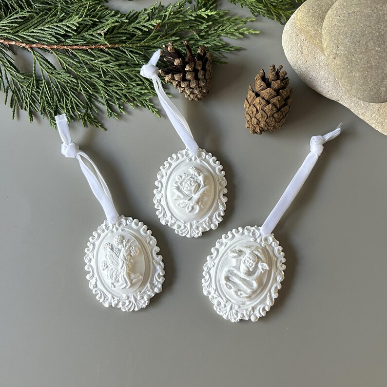 May include: Three white oval ornaments featuring intricate relief designs of a figure, a flower, and a skull with a snake. Each piece is framed by a decorative border and includes a white ribbon loop for hanging, providing a textured appearance.