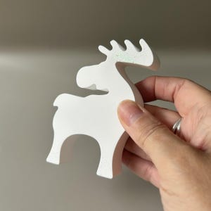 May include: A white, matte-finished moose figurine with a stylized, minimalist silhouette. The antlers feature a subtle, iridescent glitter coating that catches the light, providing a slight shimmer against the solid white surface of the object.
