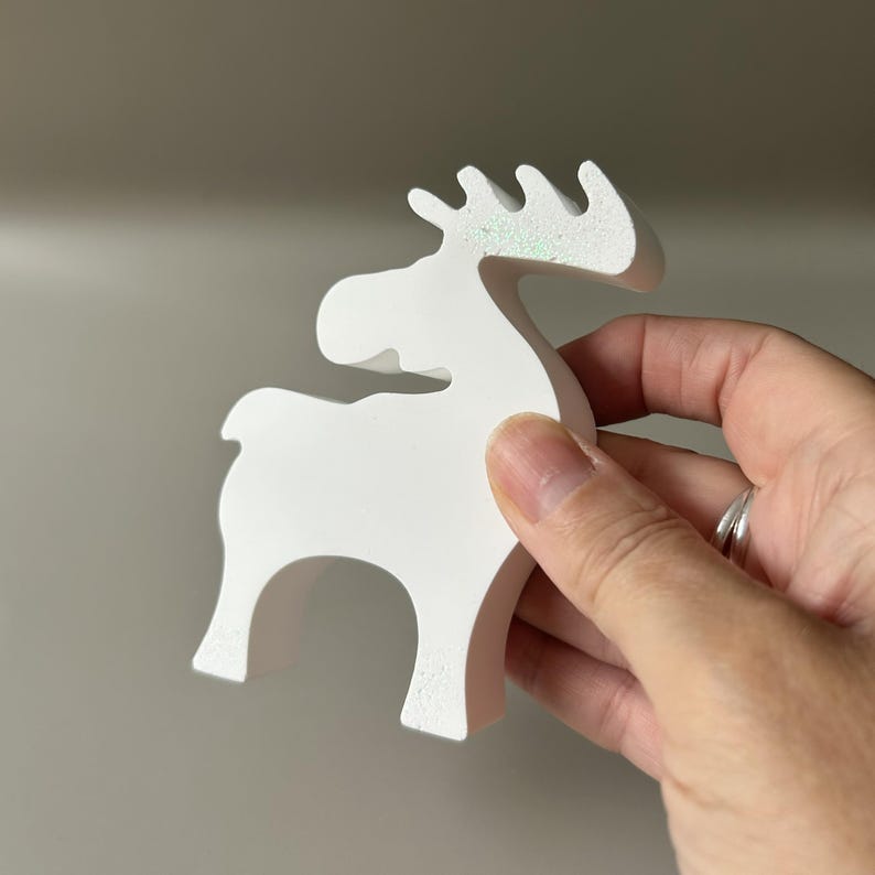 May include: A white, matte-finished moose figurine with a stylized, minimalist silhouette. The antlers feature a subtle, iridescent glitter coating that catches the light, providing a slight shimmer against the solid white surface of the object.