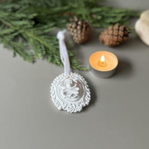K&ouml;nnte beinhalten: Ein wei&szlig;es, ovales H&auml;ngeornament mit einem Relief eines Sch&auml;dels, der von einer Schlange umschlungen ist, umgeben von einem verzierten Rand. Das Ornament ist an einem wei&szlig;en Band befestigt und weist eine matte Oberfl&auml;che auf.