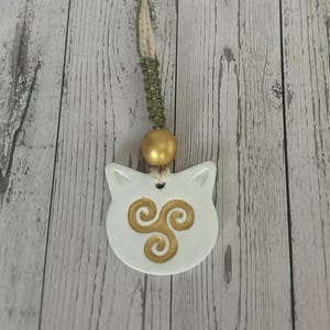May include: A white ceramic cat-shaped pendant with a gold spiral design. It hangs from a beige cord with a green macrame section and a gold bead. The background is a light wood grain.