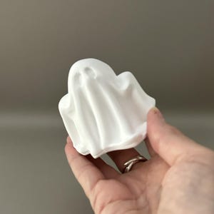 Może przedstawiać: Mała, matowa biała figurka w kształcie ducha z zaokrągloną głową i płynnymi, przypominającymi tkaninę fałdami. Obiekt posiada dwa wgłębienia w kształcie k&oacute;ł na oczy oraz gładką, jednolitą strukturę powierzchni na całej swojej formie.