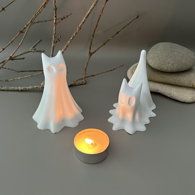 May include: Two white, matte-finished ghost figurines with pointed ears and hollow eye sockets, positioned next to a small, round, lit tea light candle. The ghosts have a smooth, flowing, draped appearance, with one standing upright and one slightly lower.