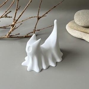 May include: A matte white ceramic figurine shaped like a stylised cat with a draped, ghost-like body. The figure features pointed ears, a smooth surface, and a tall, upright tail. The base is wide and scalloped, creating a flowing, fabric-like appearance.