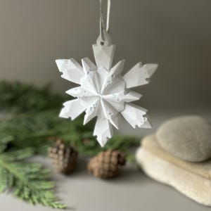 Pode incluir: Um ornamento de floco de neve branco com m&uacute;ltiplas facetas e uma estrutura geom&eacute;trica cristalina. A superf&iacute;cie apresenta manchas iridescentes subtis que refletem a luz e um acabamento mate. Est&aacute; suspenso por um cord&atilde;o fino de cor clara.