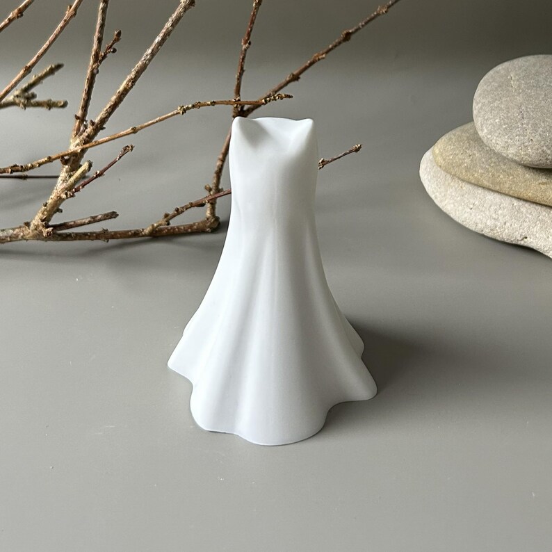 May include: A small, white, smooth ceramic figurine shaped like a flowing, flared dress or gown. The object has a solid, opaque finish with a wide, wavy base that tapers upwards to a narrow, notched top, creating a simple, sculptural form.