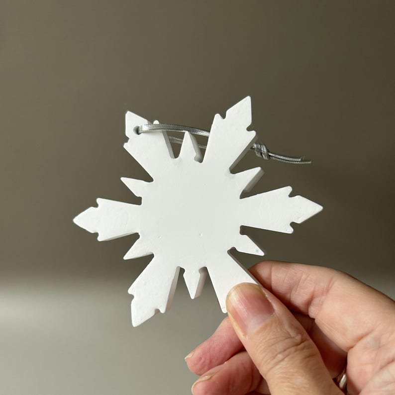 Pode incluir: Um ornamento de floco de neve branco mate com um design geom&eacute;trico de estrela de v&aacute;rias pontas. O ornamento apresenta uma superf&iacute;cie lisa e s&oacute;lida e inclui um cord&atilde;o fino de cor prateada passado pelo topo para pendurar o objeto.