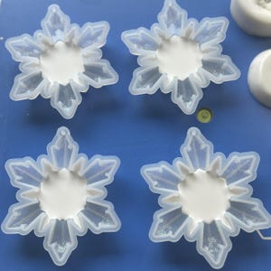 Pode incluir: Quatro moldes de silicone transl&uacute;cido em forma de flocos de neve de seis pontas dispostos sobre uma superf&iacute;cie azul. Cada molde apresenta uma depress&atilde;o circular central com bra&ccedil;os ramificados complexos para moldar flocos de neve detalhados.