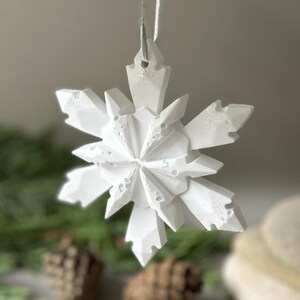 Pode incluir: Um ornamento de floco de neve branco com m&uacute;ltiplas facetas, acabamento mate e detalhes subtis de purpurina iridescente. A decora&ccedil;&atilde;o geom&eacute;trica em forma de estrela apresenta pontas angulares e est&aacute; suspensa por uma fita fina e clara.