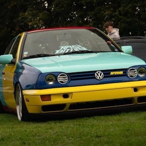 Peut inclure: Une Volkswagen Golf multicolore. Le toit est rouge, le capot bleu clair, l'avant bleu foncé et le pare-chocs jaune. Le texte "RASS BITIME" est visible sur le pare-brise.
