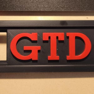 Könnte beinhalten: Schwarzes rechteckiges Emblem mit den roten Buchstaben "GTD". Die Buchstaben sind erhaben und heben sich vom schwarzen Hintergrund ab. Das Emblem ist wahrscheinlich für ein Fahrzeug.