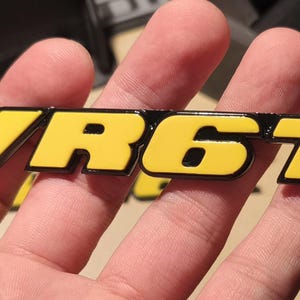 May include: A yellow and black plastic emblem with the text "VR6T" in black letters.