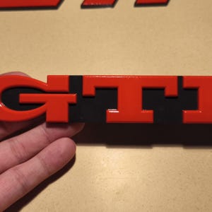 Peut inclure: Emblème GTI en plastique rouge et noir. Les lettres sont en relief et ont un contour noir.