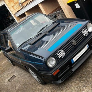 Peut inclure: Une Volkswagen Golf bleue avec un pare-chocs avant noir et une bande bleue et blanche sur le capot. La voiture a deux phares Hella et un logo Volkswagen sur la calandre.