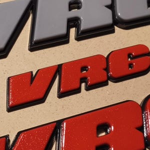May include: Close-up of a red and black "WRC" logo. The letters are raised and have a textured surface.