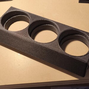 Peut inclure: Un support gris foncé imprimé en 3D avec trois ouvertures circulaires. La base rectangulaire a une surface texturée. Les ouvertures sont conçues pour contenir des objets cylindriques. Le support est en matériau solide et durable.