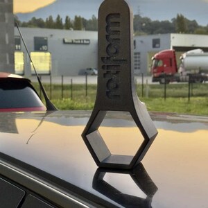 Op de afbeelding: Een zwart, sleutelvormig object met het woord "rotiform" in verhoogde letters. Het object rust op het dak van een auto, met een achtergrond van gebouwen en een zonsondergang.