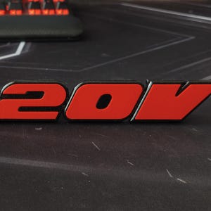 Peut inclure: Un emblème rouge et noir avec le texte "20V". Les chiffres et la lettre sont en gras, en police sans-serif. L'emblème est probablement destiné à un véhicule ou une machine.