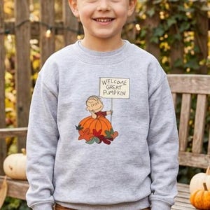 Peut inclure: Un sweat-shirt gris clair à col rond avec une impression graphique représentant un personnage de dessin animé assis sur une grosse citrouille orange. Le personnage tient un panneau indiquant "WELCOME GREAT PUMPKIN" entouré de feuilles d'automne.