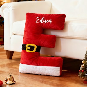 Puede incluir: Un gran cojín rojo de felpa con forma de letra "E" con cinturón negro y hebilla dorada, y ribete blanco. El nombre "Edison" está escrito en el cojín. Un pequeño árbol de Navidad, un calcetín y una campana están en el suelo de madera.