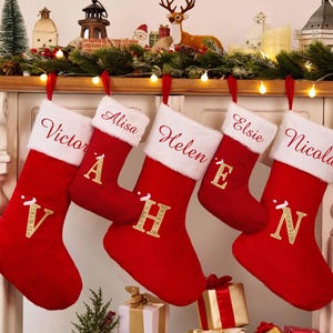 Può includere: Cinque calze di Natale rosse con polsini in finta pelliccia bianca sono appese sopra un camino, ognuna personalizzata con un nome in corsivo e un'iniziale dorata. Le decorazioni festive includono ghirlande, luci e un Babbo Natale.