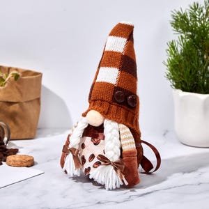 May include: A coffee-themed gnome doll with a brown and white checkered hat and a white beard. The gnome's outfit features a coffee bean pattern and brown accents. The doll is decorated with two brown buttons and a small bow.