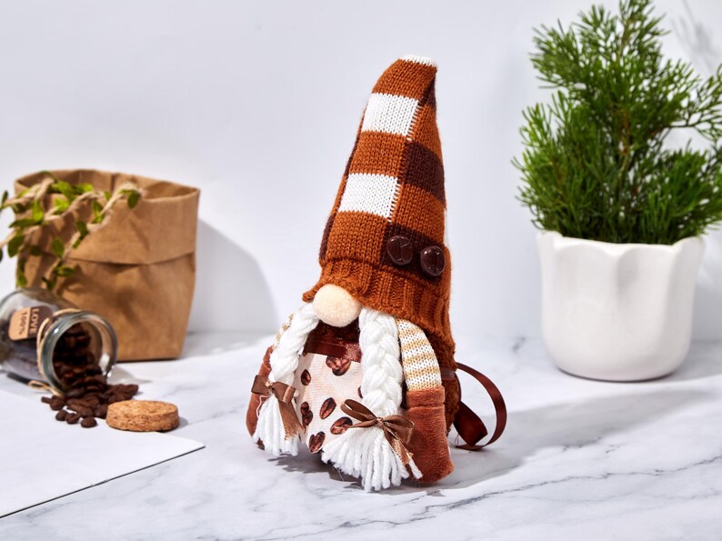 May include: A coffee-themed gnome doll with a brown and white checkered hat and a white beard. The gnome's outfit features a coffee bean pattern and brown accents. The doll is decorated with two brown buttons and a small bow.
