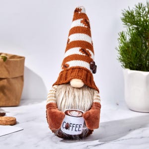 May include: A coffee-themed gnome figurine with a long, striped hat in brown and white. The gnome has a long, white beard and holds a mug that says "COFFEE". The gnome is wearing a brown sweater and mittens.
