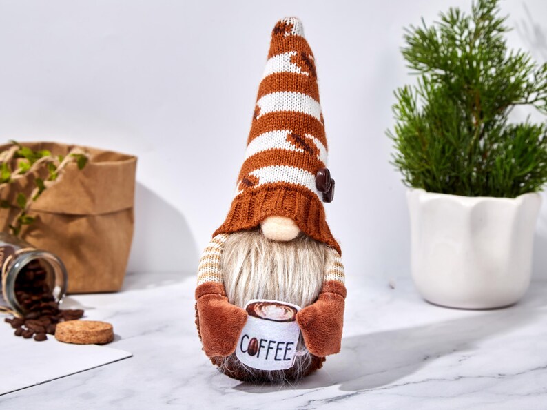 May include: A coffee-themed gnome figurine with a long, striped hat in brown and white. The gnome has a long, white beard and holds a mug that says "COFFEE". The gnome is wearing a brown sweater and mittens.