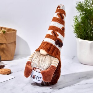 May include: A coffee-themed gnome figurine with a long, striped hat featuring coffee bean designs. The gnome has a long, white beard and holds a mug that says "COFFEE". The figurine is brown, white, and tan.