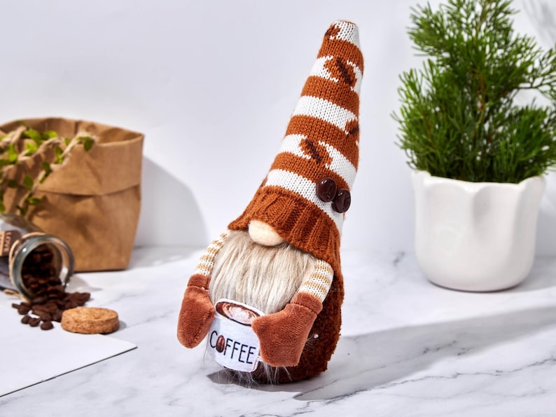 May include: A coffee-themed gnome figurine with a long, striped hat featuring coffee bean designs. The gnome has a long, white beard and holds a mug that says "COFFEE". The figurine is brown, white, and tan.